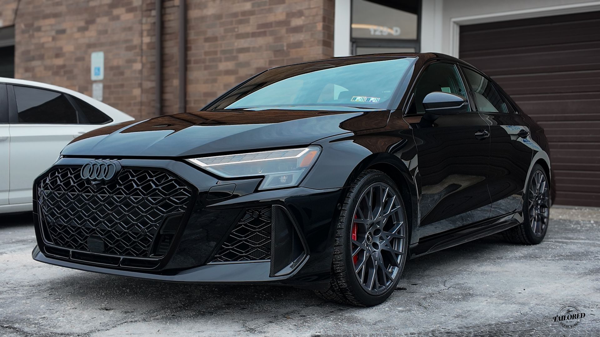 Black Audi coupe parked on a snowy driveway in front of a brick house and garage.