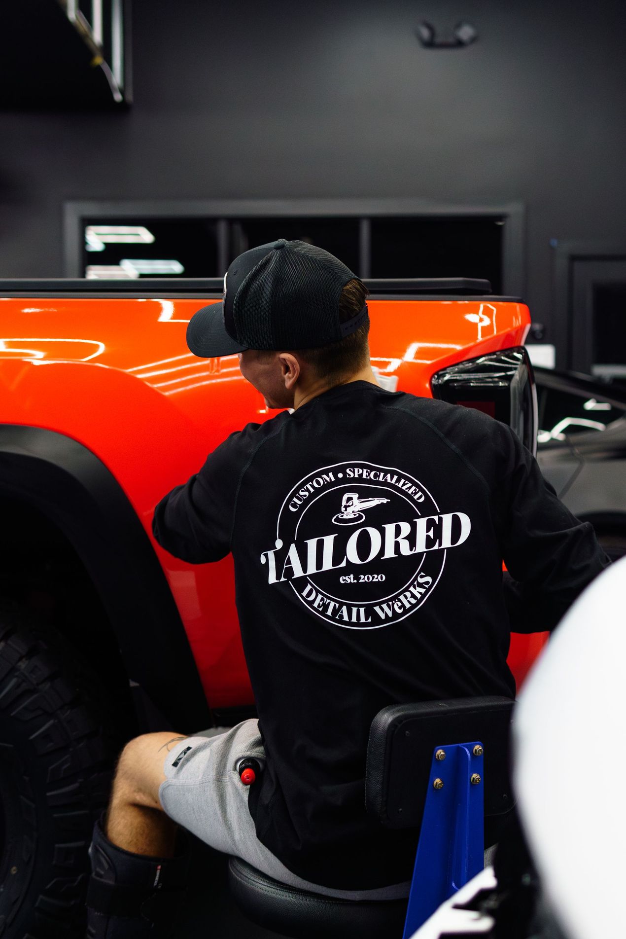 Person seated beside a bright orange car in a dark garage, wearing a black “Tailored” shirt.