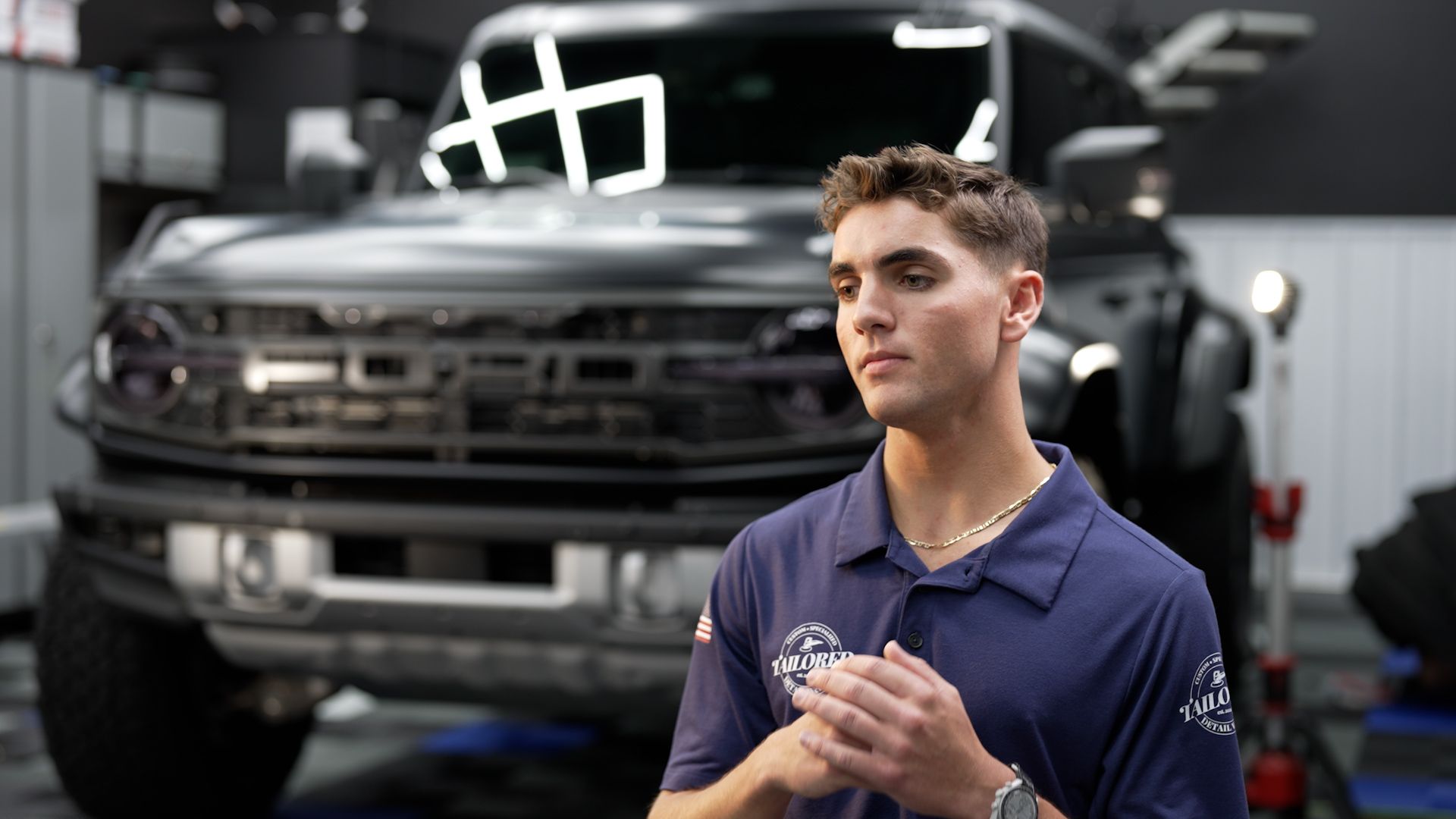 Young man in a blue shirt standing in front of a lifted truck in a garage, hands clasped