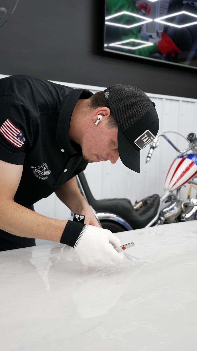 Mechanic in black polo and gloves working on a car, leaning over a white vehicle in a garage