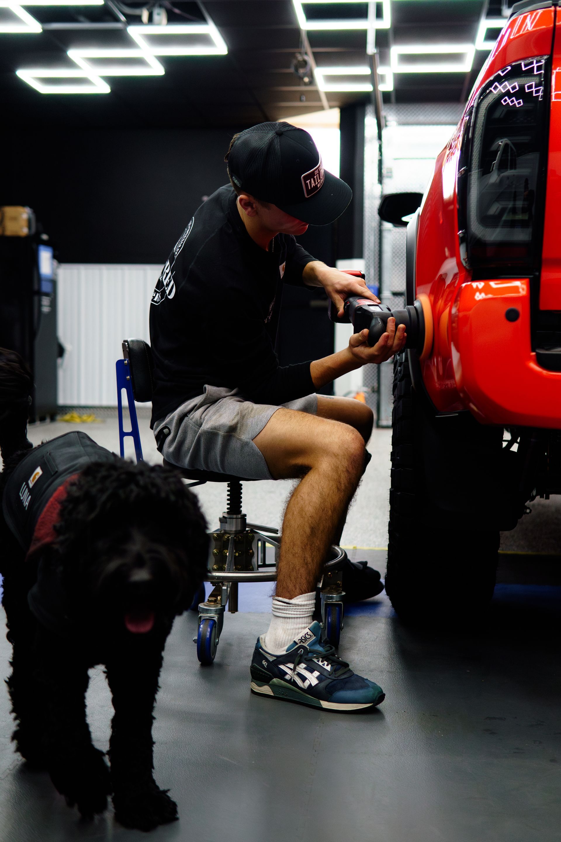 Person kneeling beside a bright red vehicle, working on it in a garage, with a black dog in the foreground