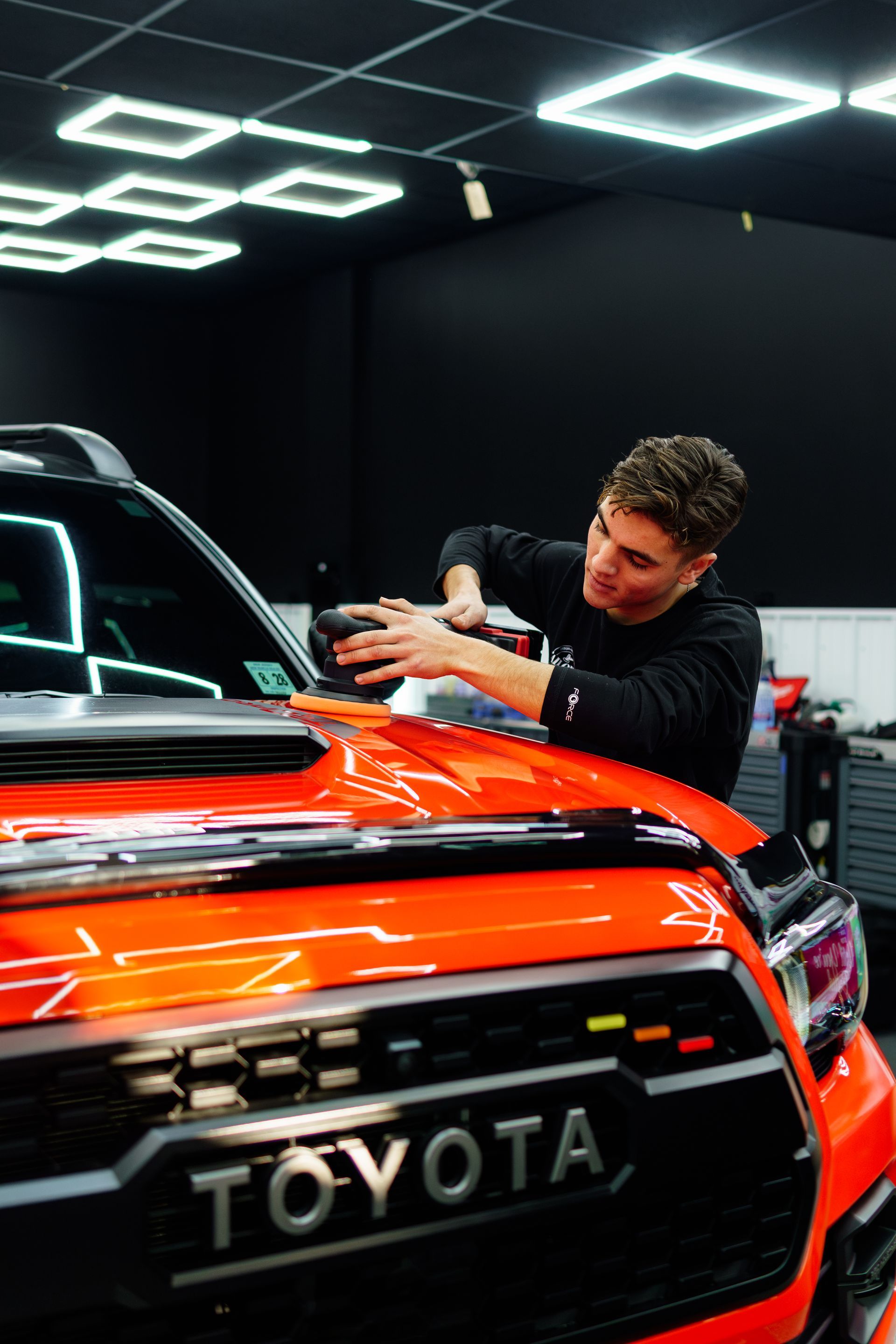 Mechanic polishing an orange Toyota in a dark studio with bright ceiling lights