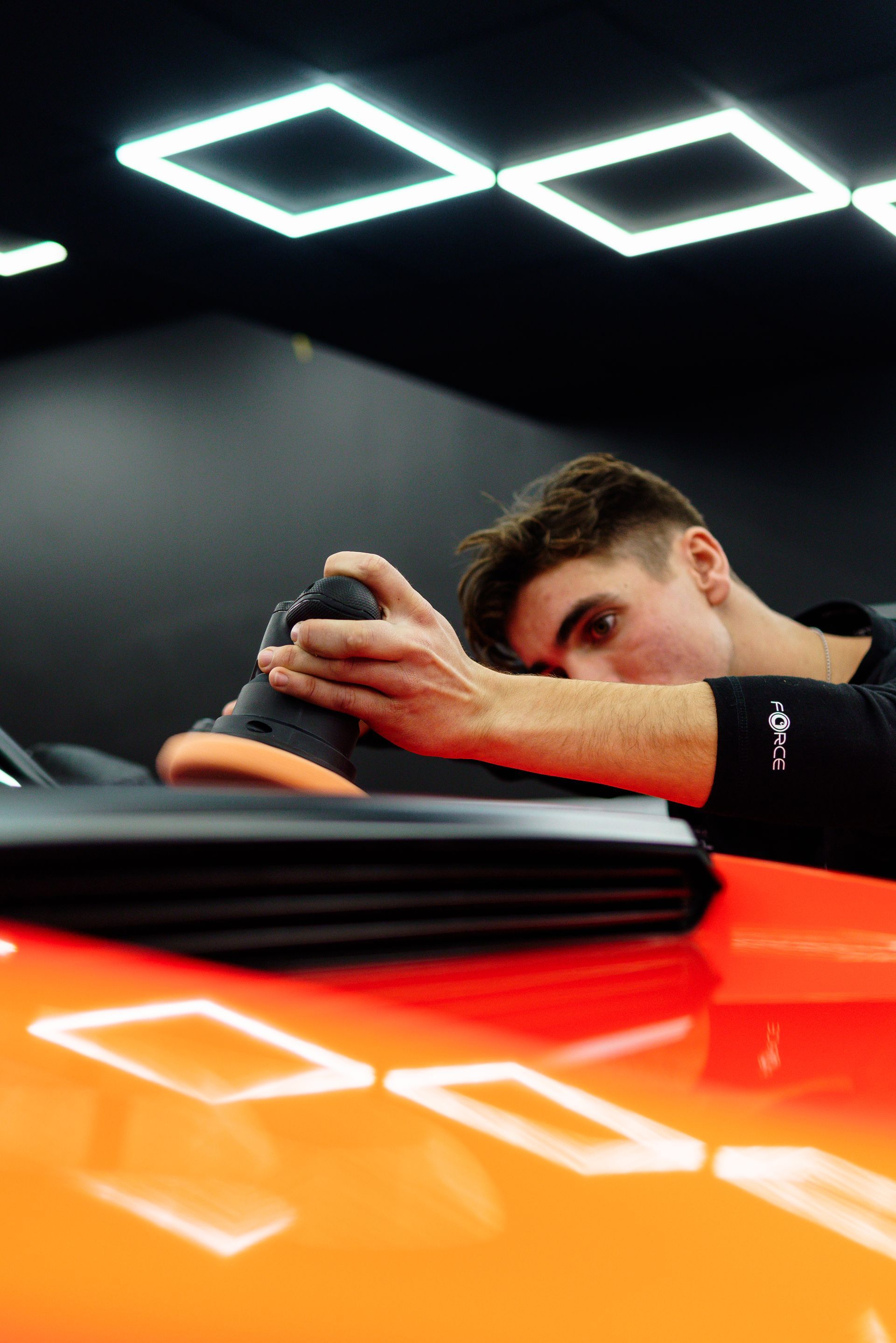 Person leaning over an orange sports car, resting hands on the roof in a dark showroom under geometric lights