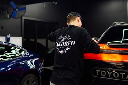 Person in a black shirt working on an orange Toyota in a garage next to a dark blue car