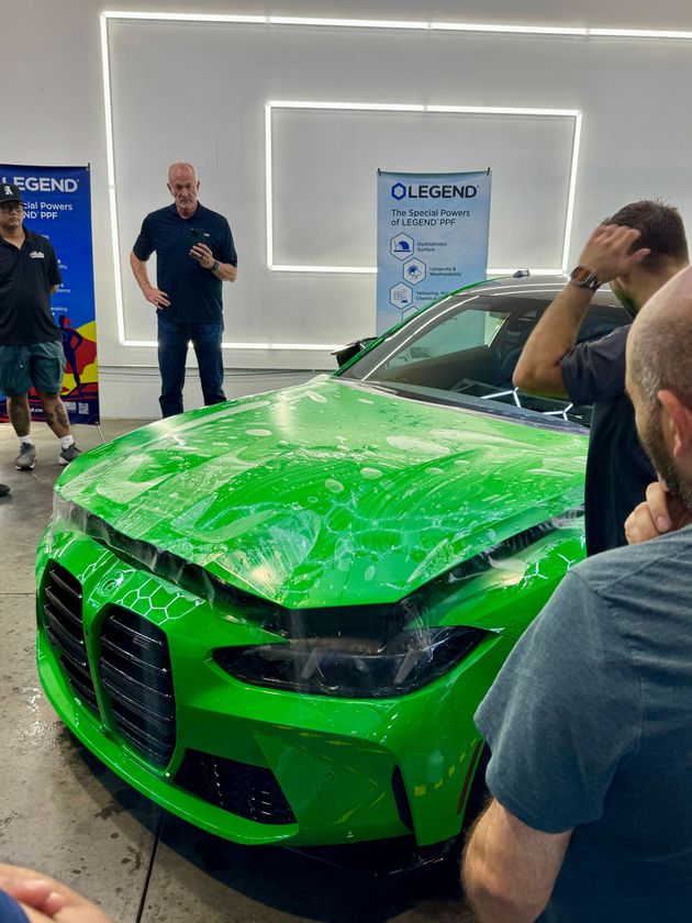 Bright green BMW-style sports car with shattered windshield at an indoor event, people standing around it