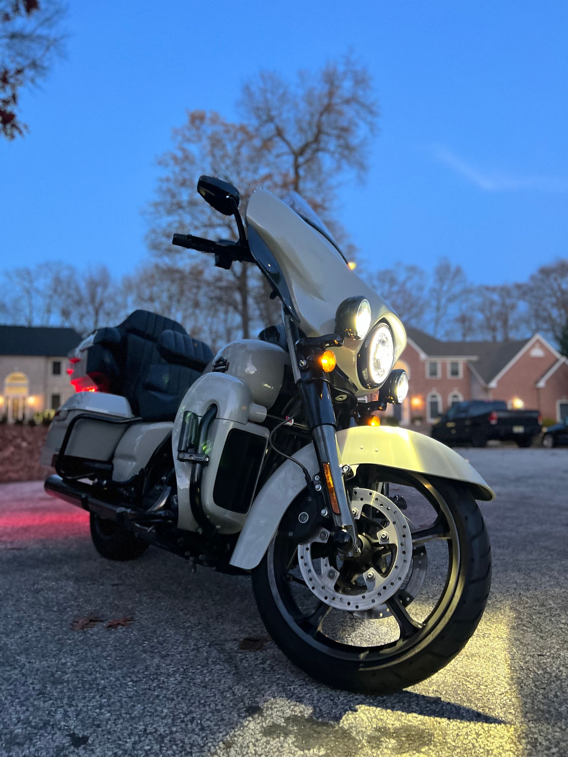 White touring motorcycle parked on wet pavement at dusk, with headlight on in a residential street.