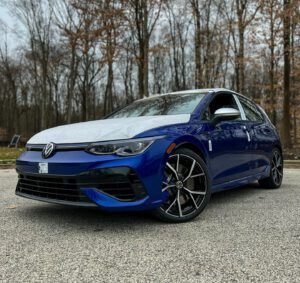 Blue Volkswagen hatchback parked on gravel with bare trees in the background