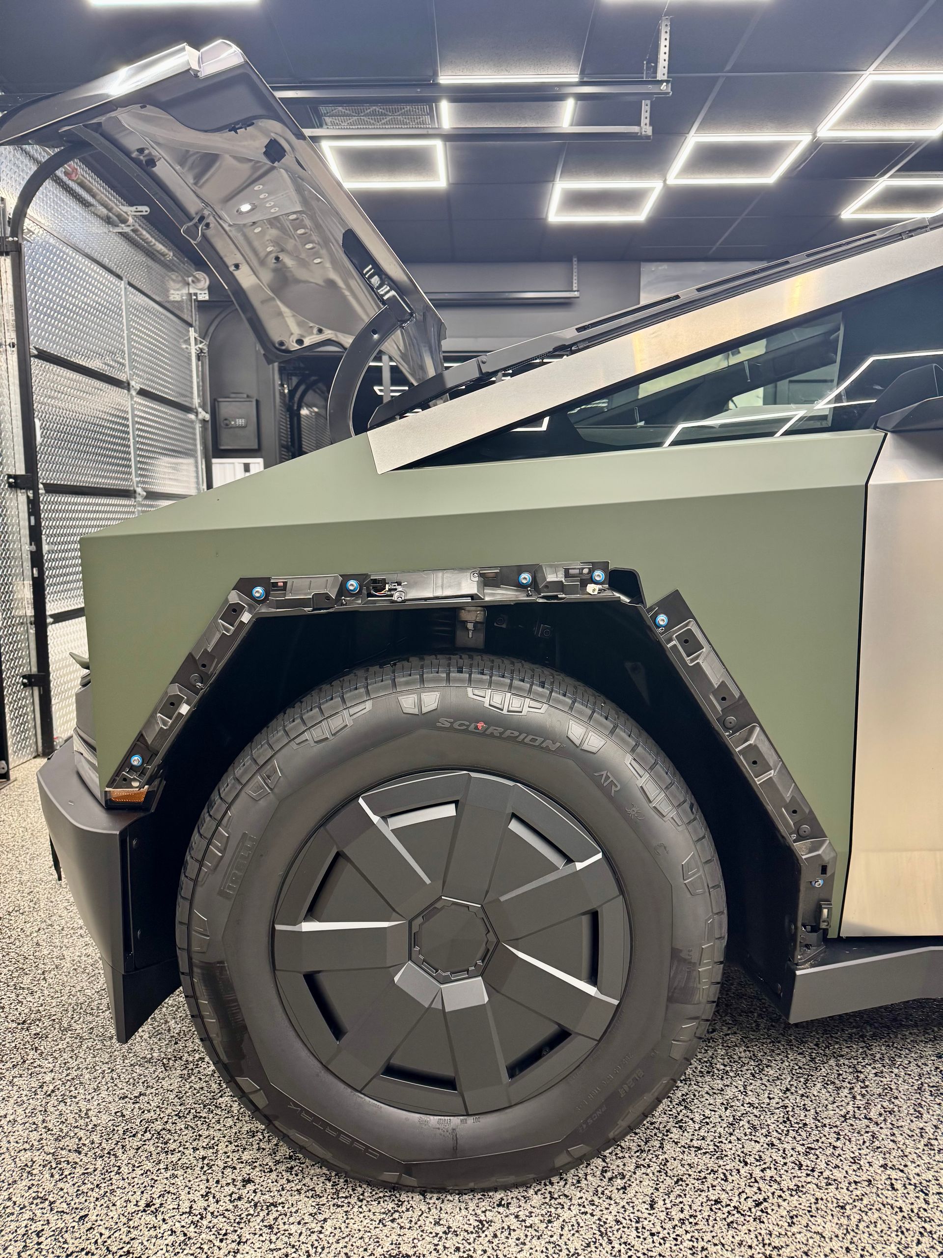 Close-up of a matte olive-green armored vehicle’s rear wheel and fender in a showroom or garage.