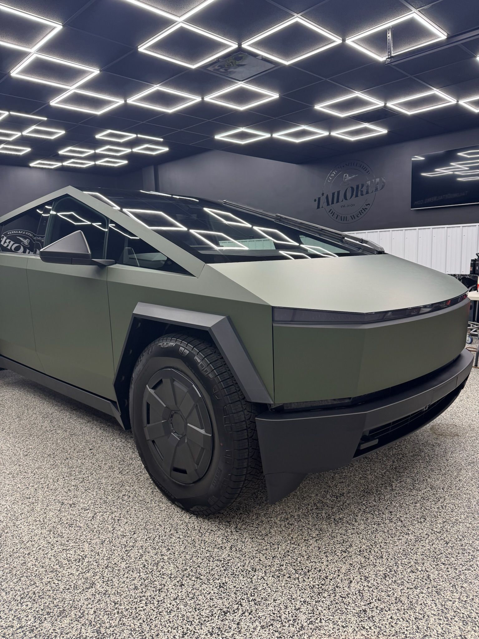 Matte olive-green Cybertruck in a showroom under geometric ceiling lights