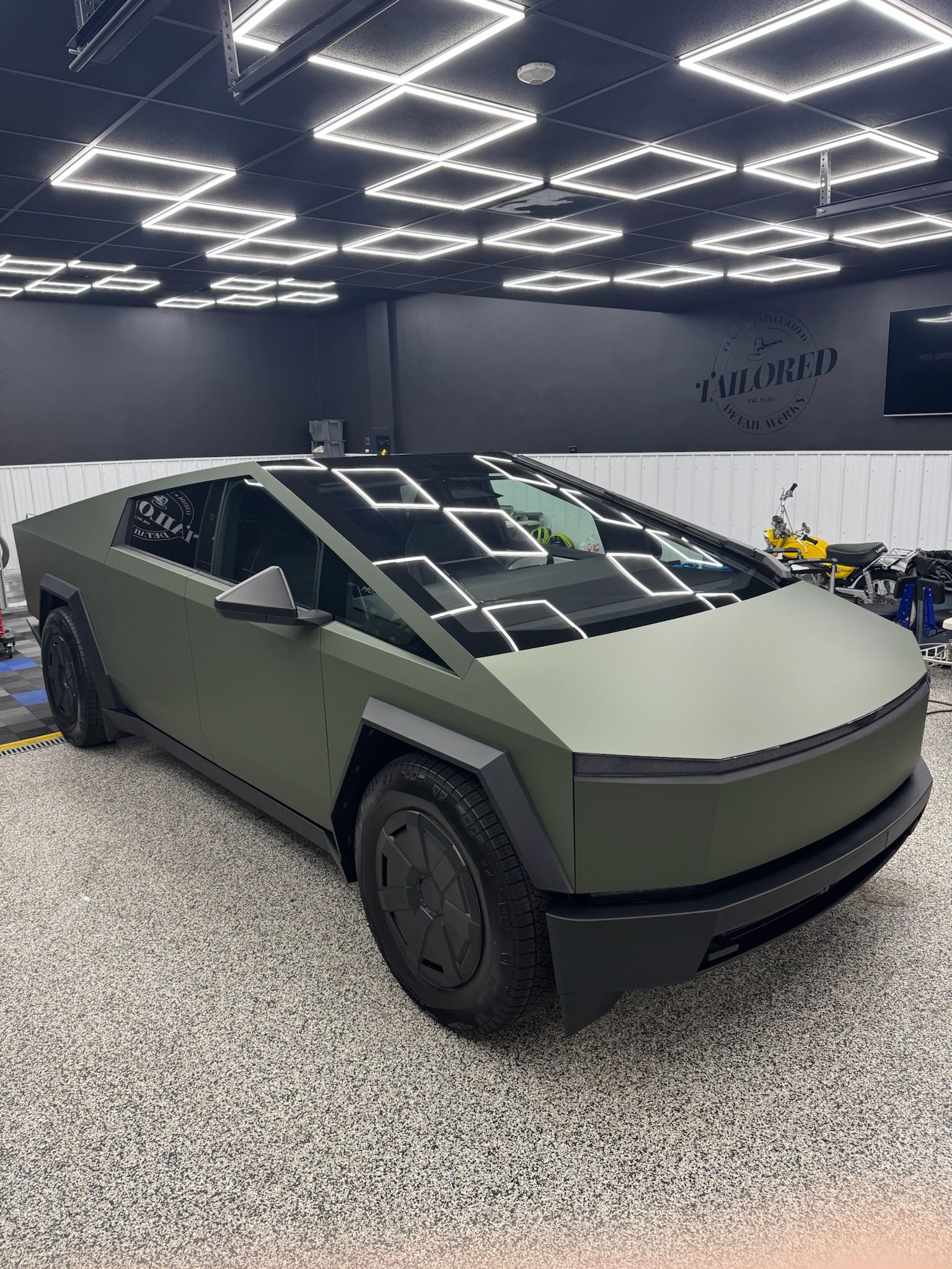 Matte olive Cybertruck in a showroom with geometric ceiling lights and gravel floor