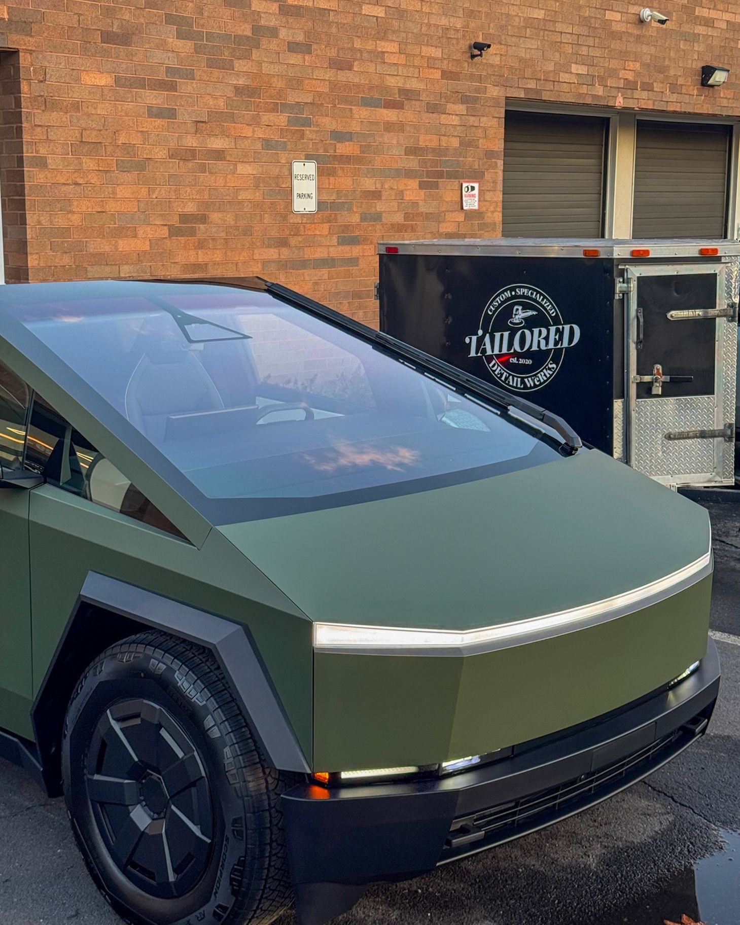 Olive-green Tesla Cybertruck parked outside a brick building beside a trailer