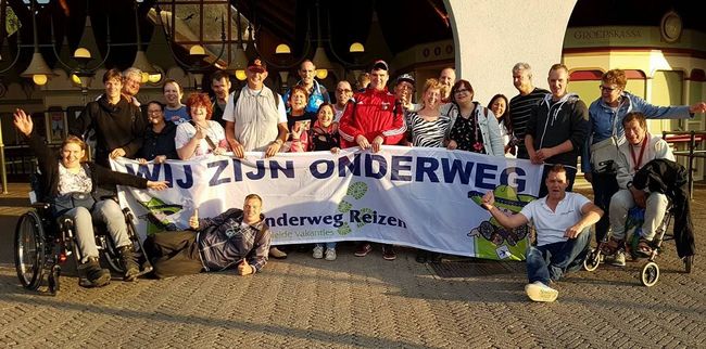 Een groep poseert buiten achter een spandoek, waarbij sommigen in een rolstoel zitten onder fel licht.