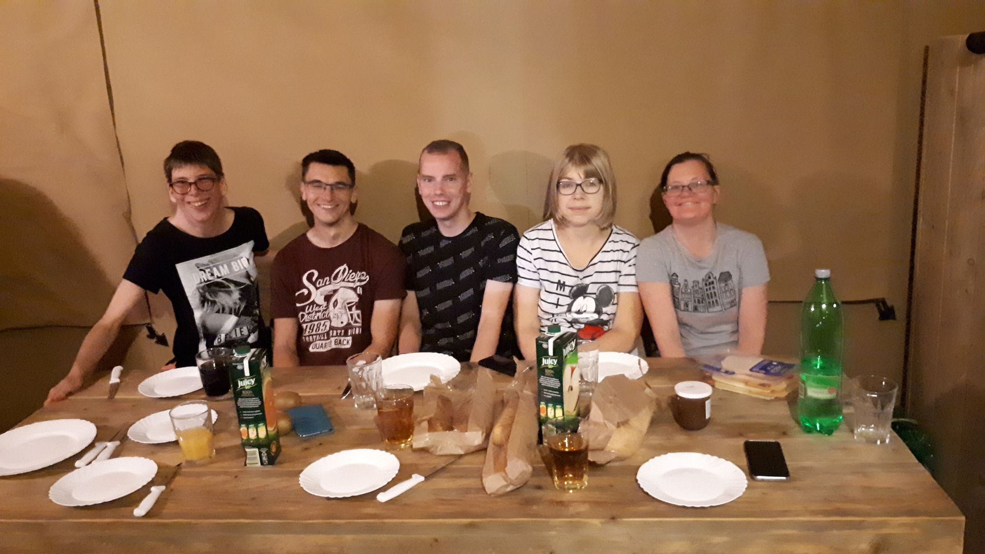Vijf personen zitten aan een houten tafel met borden en drankjes in een eenvoudige binnenruimte.