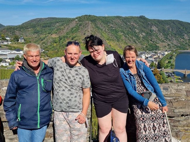 Vier mensen poseren buiten met heuvels en water op de achtergrond, lachend op een zonnige dag.