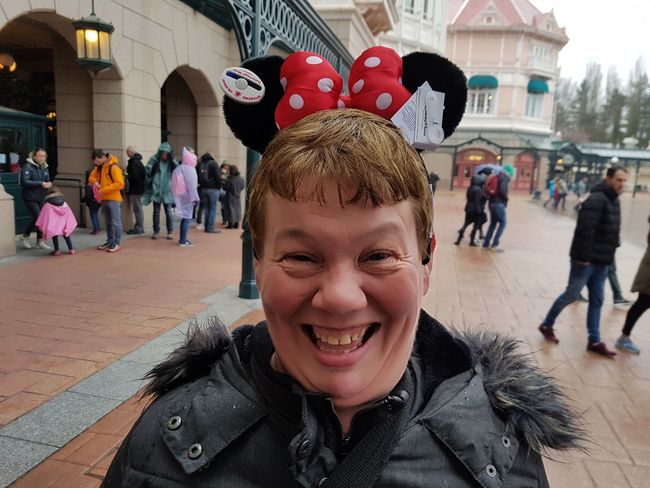 Een lachend persoon met Minnie Mouse-oren in een pretpark, met bezoekers en gebouwen op de achtergrond.