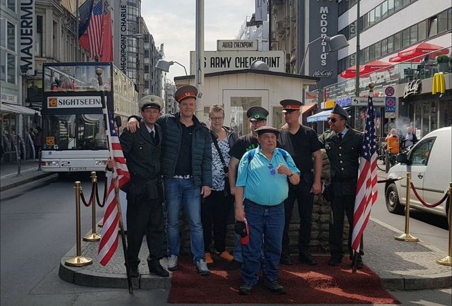 Een groep poseert op een stadsstoep met een rode loper, gestreepte lantaarnpalen en drukke winkels op de achtergrond.
