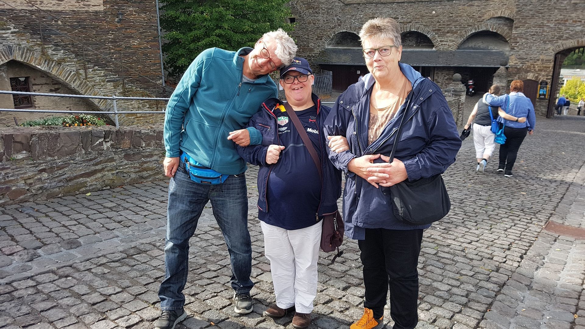 Drie mensen poseren op een geplaveide binnenplaats, met oude bakstenen muren op de achtergrond.