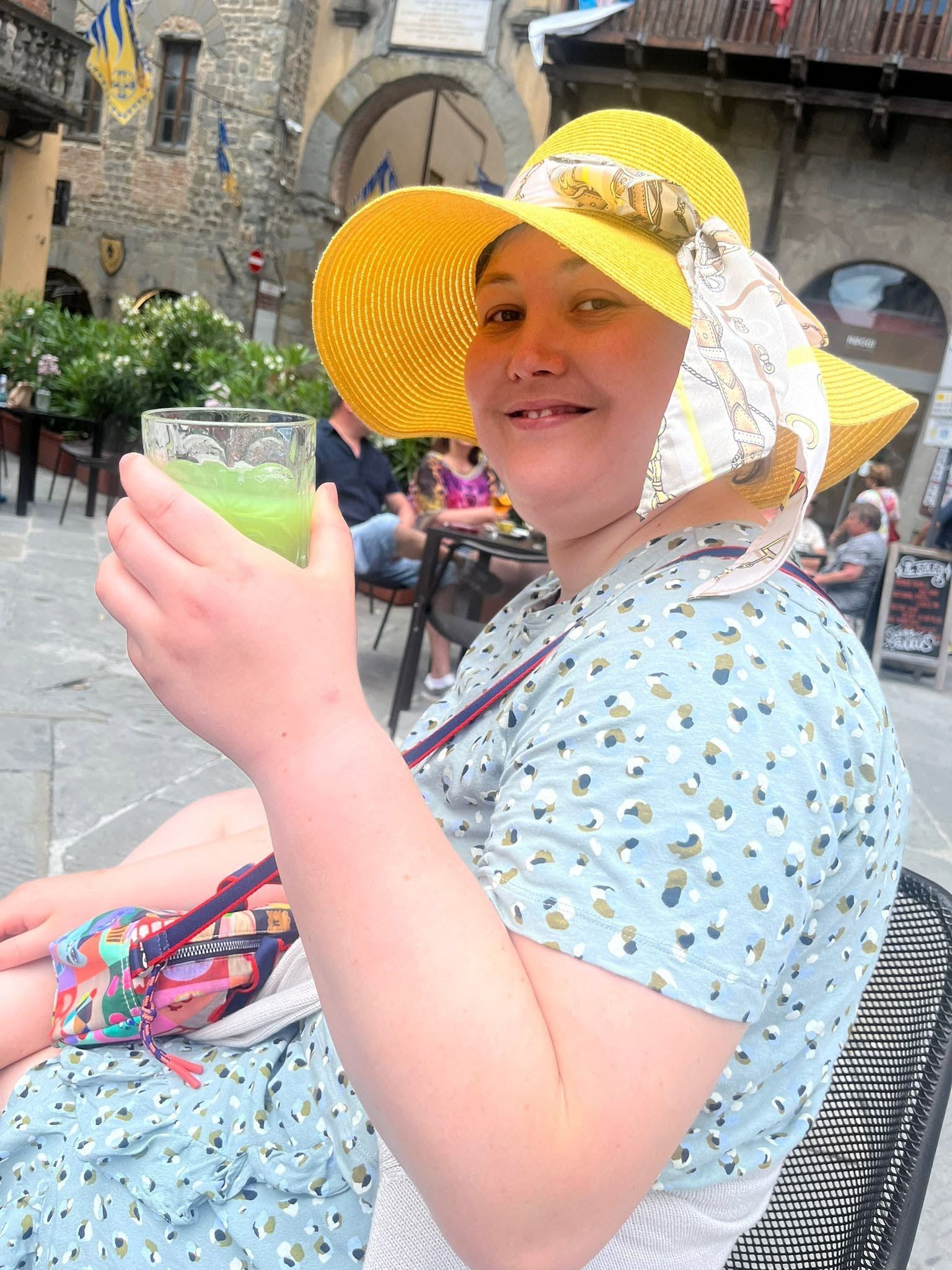 Vrouw met een gele zonnehoed en een groen drankje in haar hand, zittend op een stenen binnenplaats.