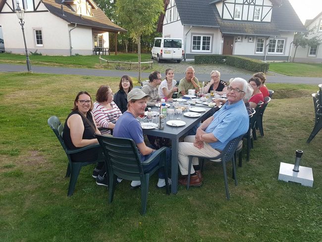 Mensen zitten rond een lange eettafel in een grasveld, met huizen op de achtergrond.