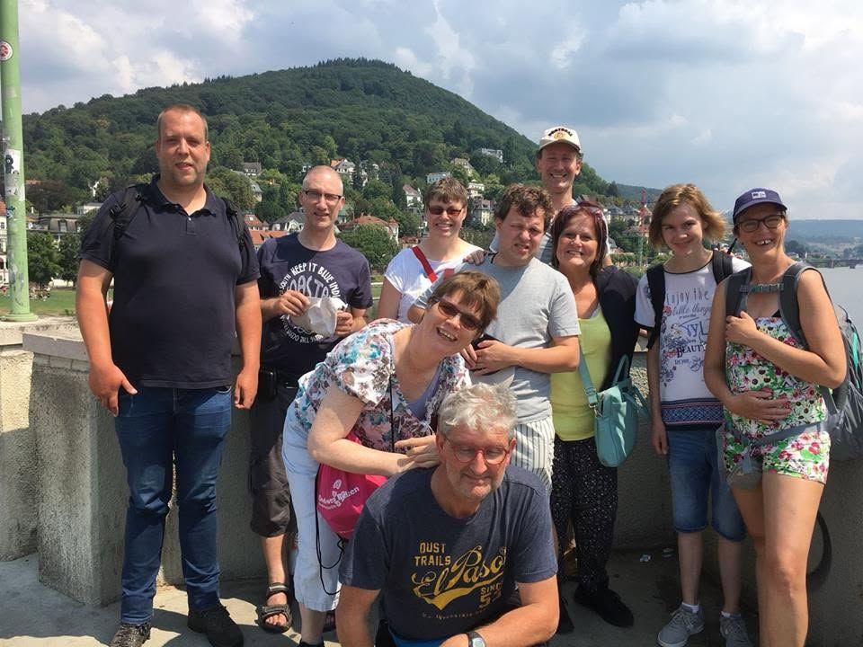 Een groep mensen poseert buiten bij een heuvel en water, en lacht naar de camera.
