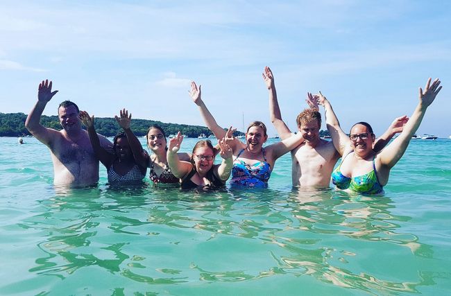 Een groep mensen steekt hun armen omhoog terwijl ze in het turquoise oceaanwater bij een strand drijven.
