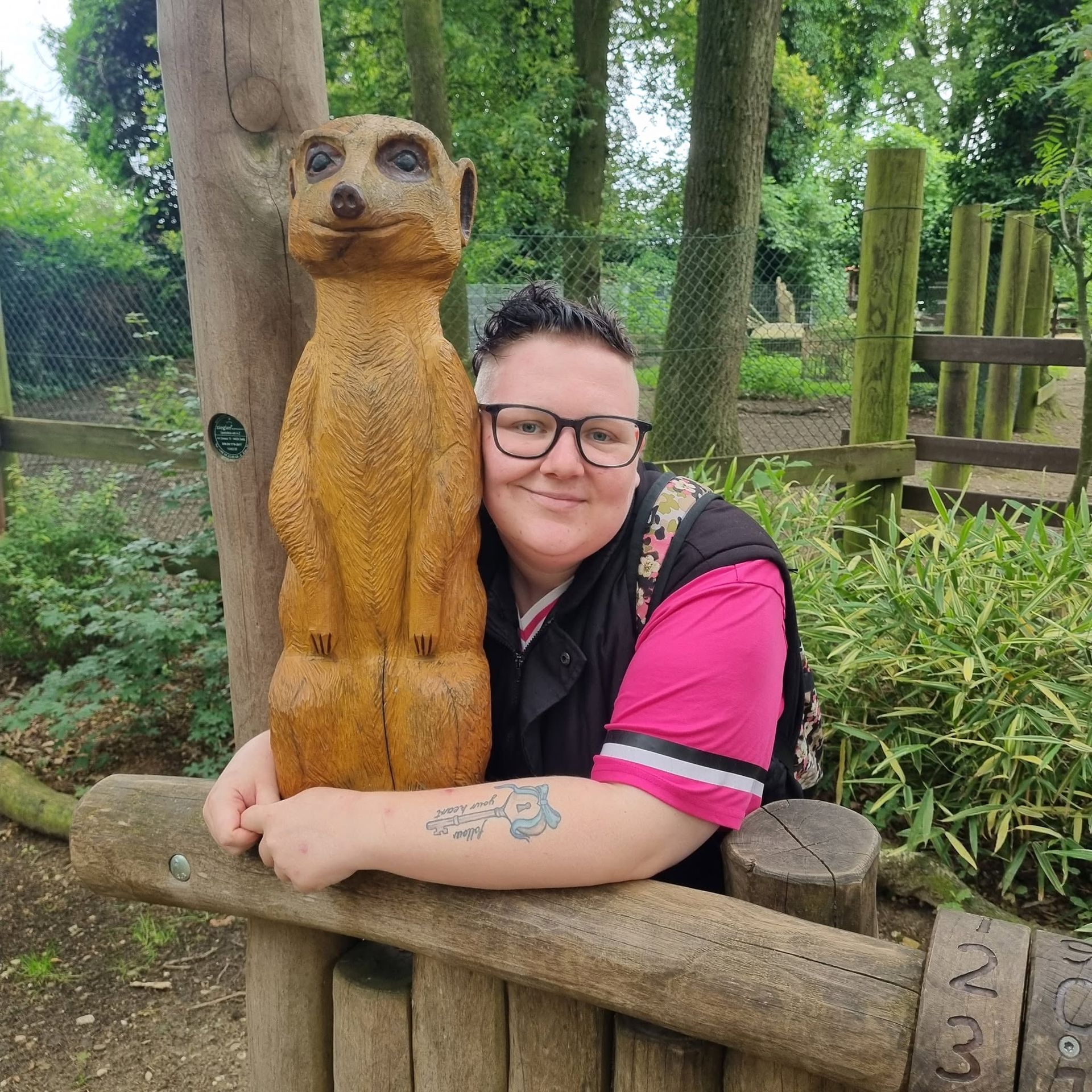 Een persoon omhelst een gebeeldhouwd dierenbeeld in een lommerrijk park en glimlacht achter een bril.