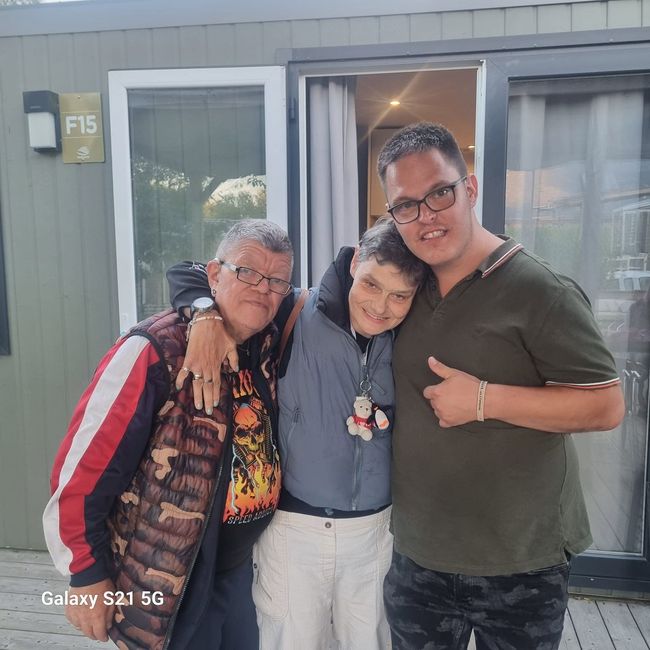 Drie mensen staan ​​lachend en elkaar omhelzend voor een deuropening, waarvan er één een duim omhoog steekt.