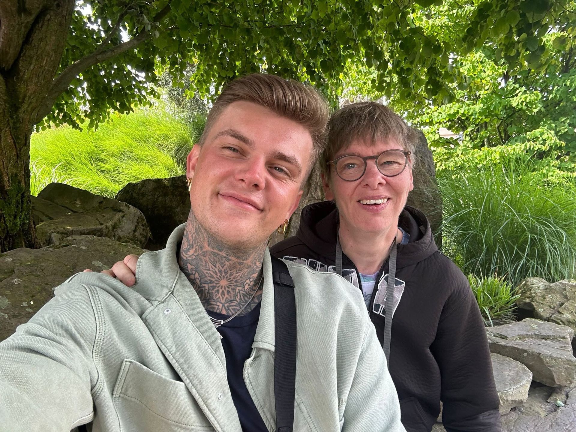 Twee mensen glimlachen onder een boom met veel bladeren naast grote rotsen, waarvan er één een selfie maakt.