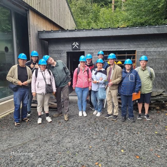 Een groep mensen met blauwe veiligheidshelmen verzamelde zich buiten een mijningang en poseerde op een grindterrein.