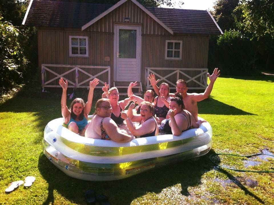 Mensen spetteren in een klein opblaasbaar zwembad op een zonnig gazon voor een houten hut.