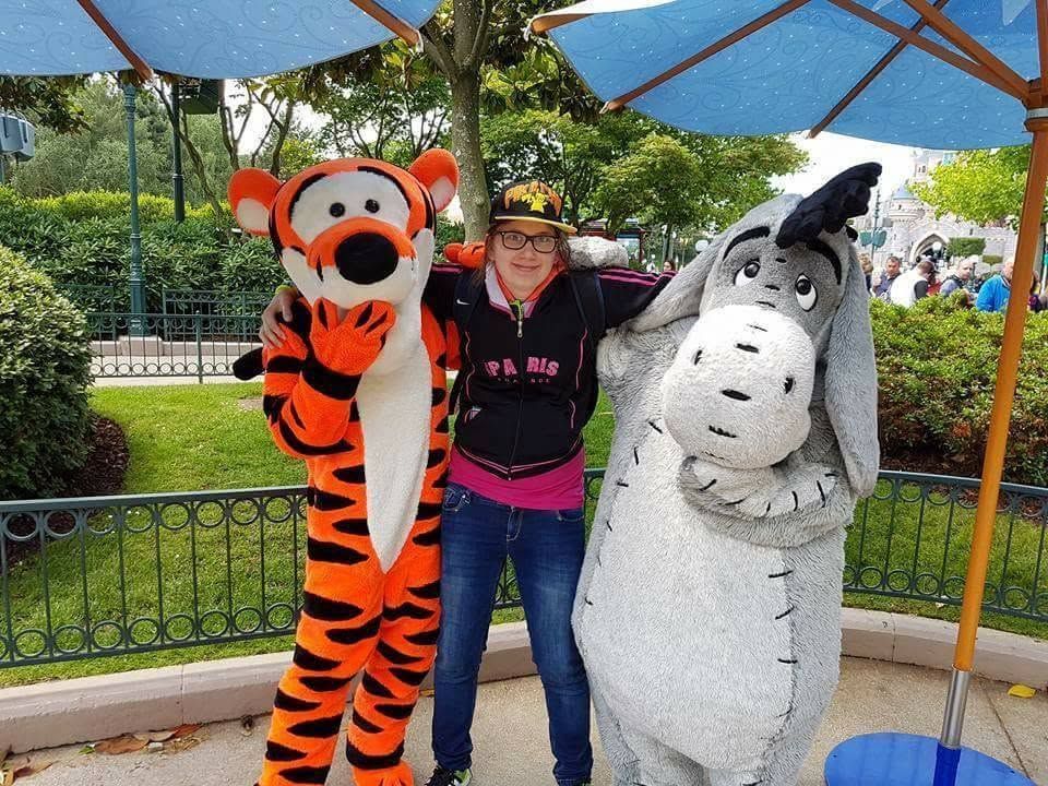 Een persoon poseert met de figuren van Tigger en Eeyore onder een blauwe luifel in een park.