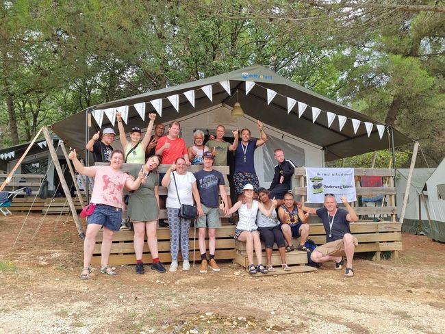 Een groep mensen poseert voor een versierde tent, lachend en zwaaiend in de buitenlucht onder de bomen.