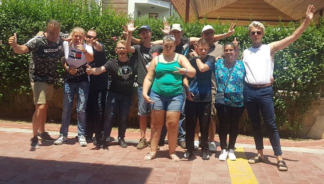Een groep mensen poseert buiten voor een heg, sommigen glimlachen en steken hun armen omhoog.