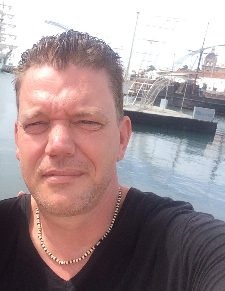 Selfie van een man in een zwart shirt en met een gouden ketting in een jachthaven met boten op de achtergrond.