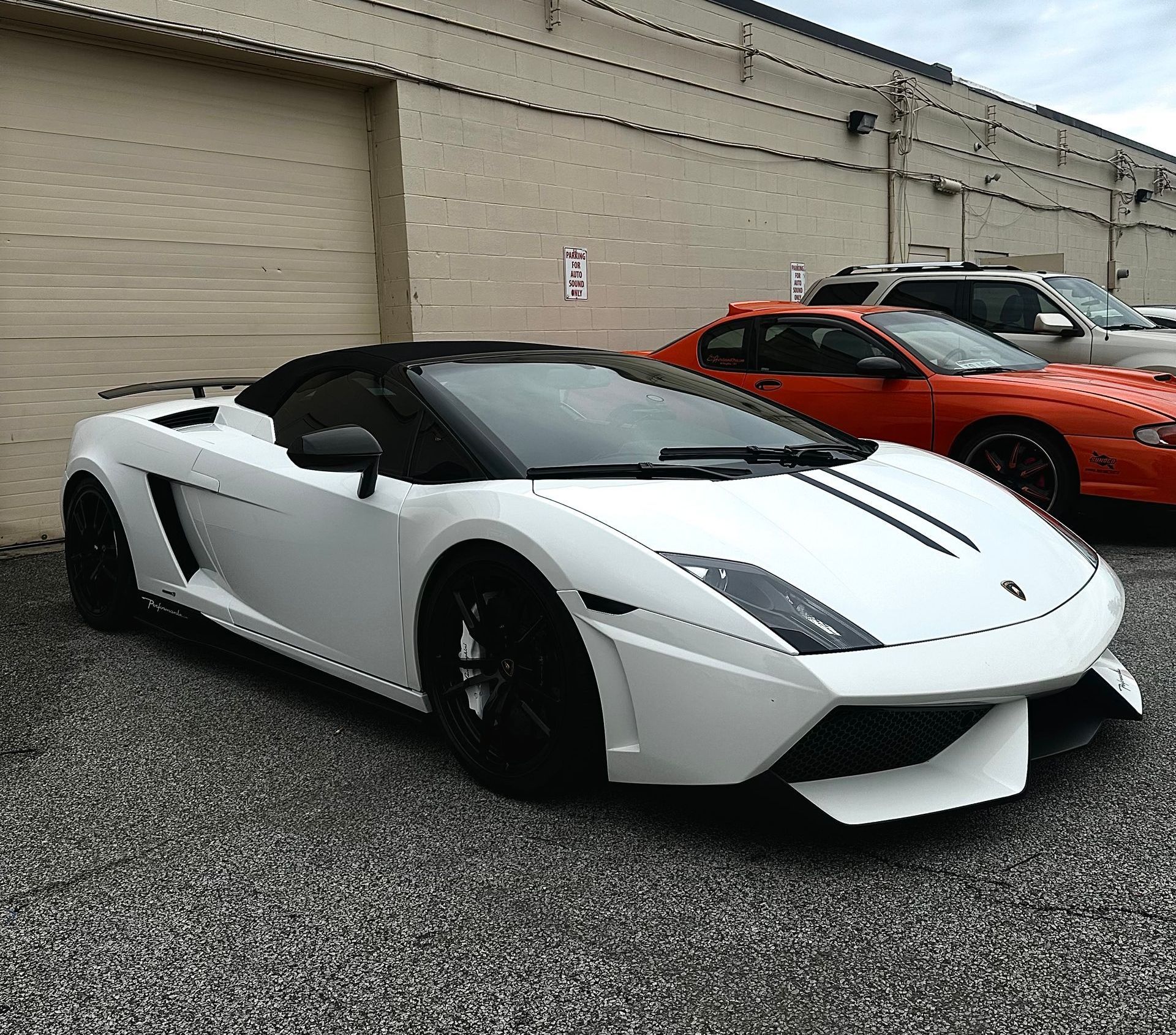 White Lamborghini coupe parked on gravel beside an orange sports car in front of a beige building