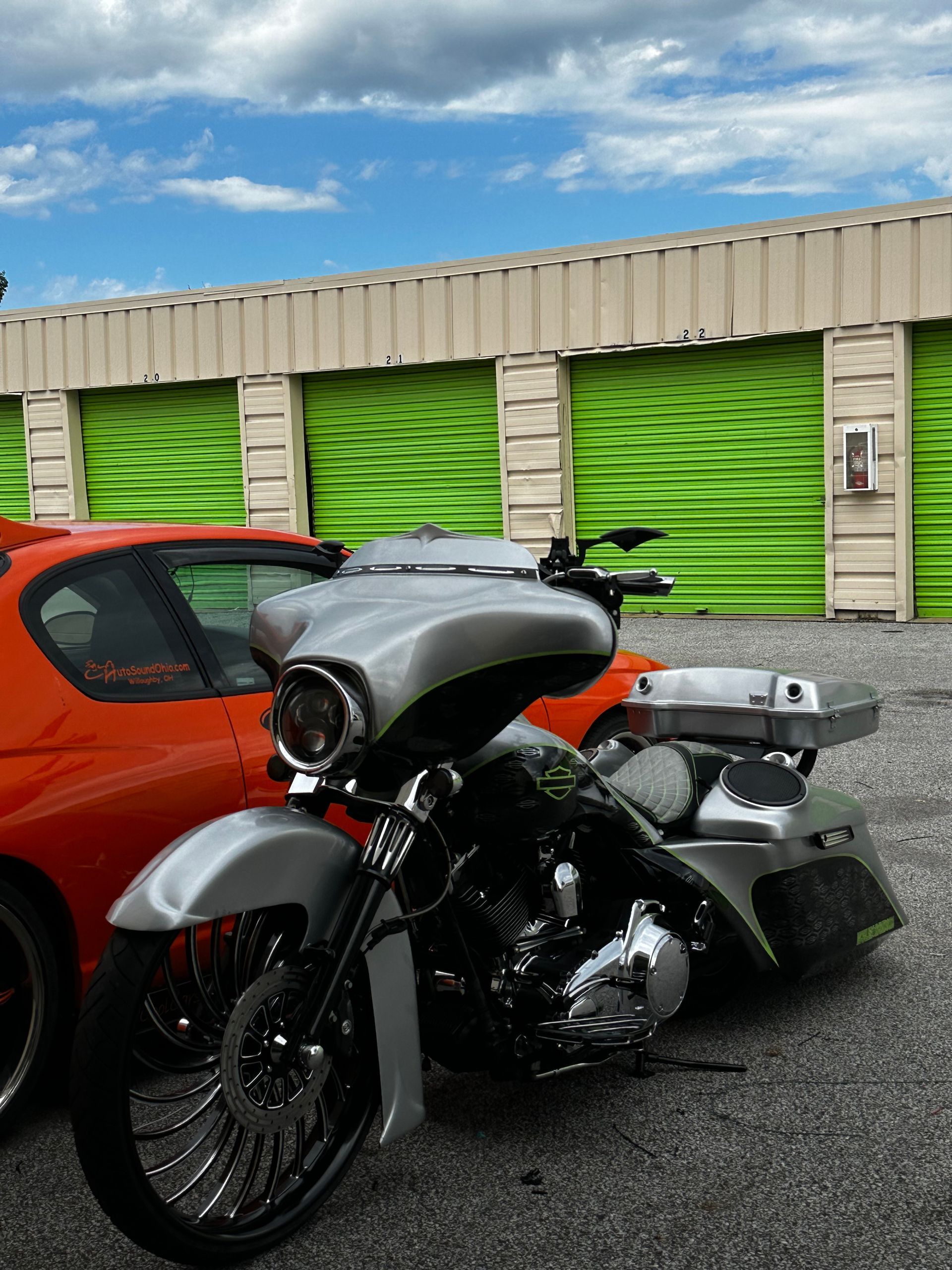 Silver motorcycle parked beside an orange car in front of green storage units under a blue sky.