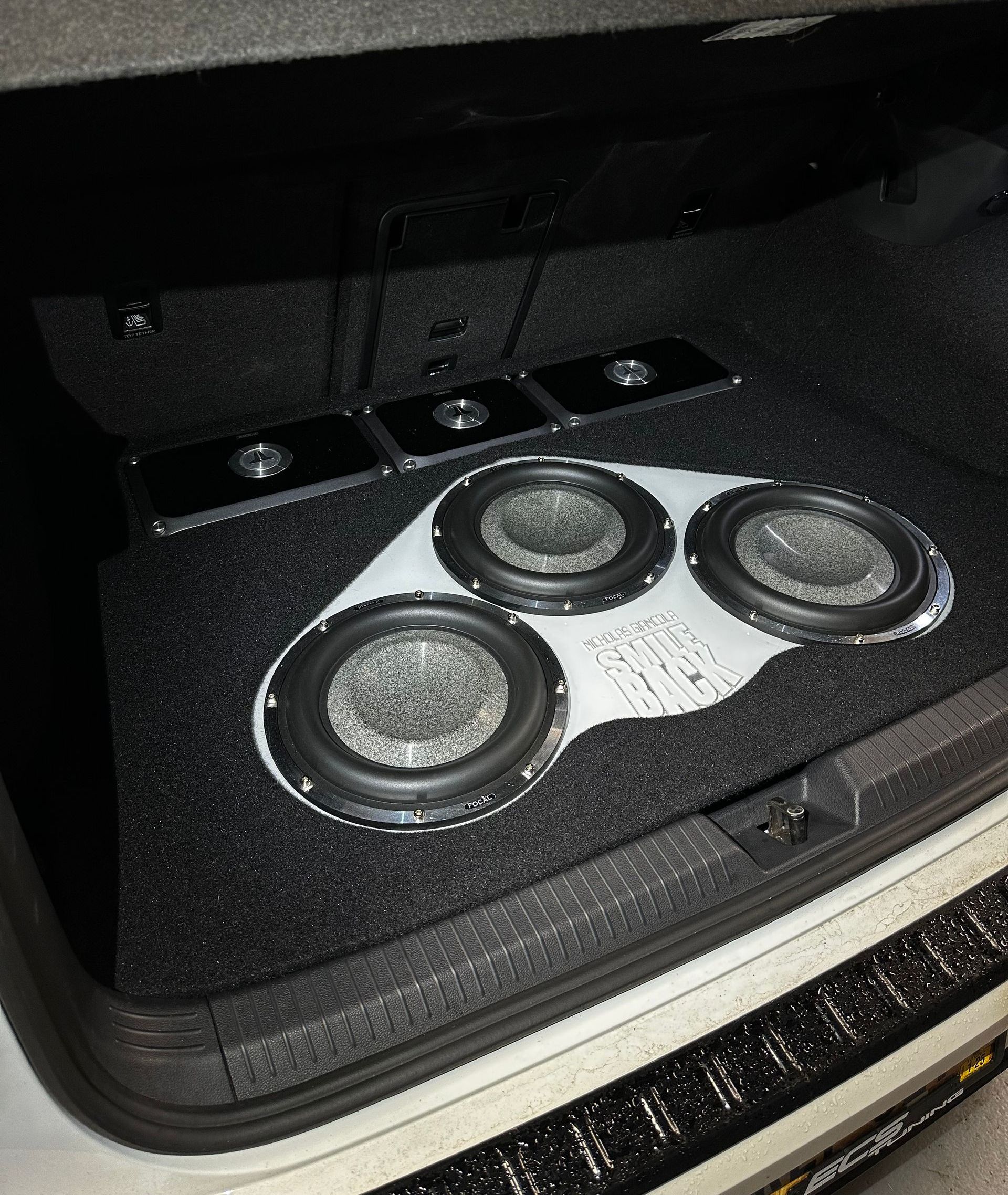 Three speakers mounted in a car trunk subwoofer enclosure with black and silver trim