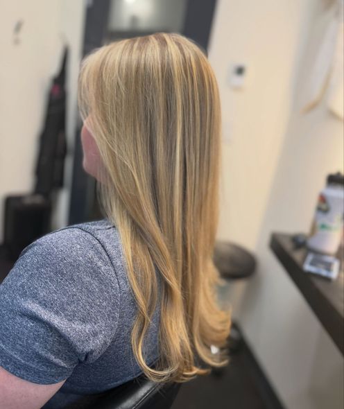 Woman with long blonde hair in a salon chair, shown from the side wearing a gray shirt.