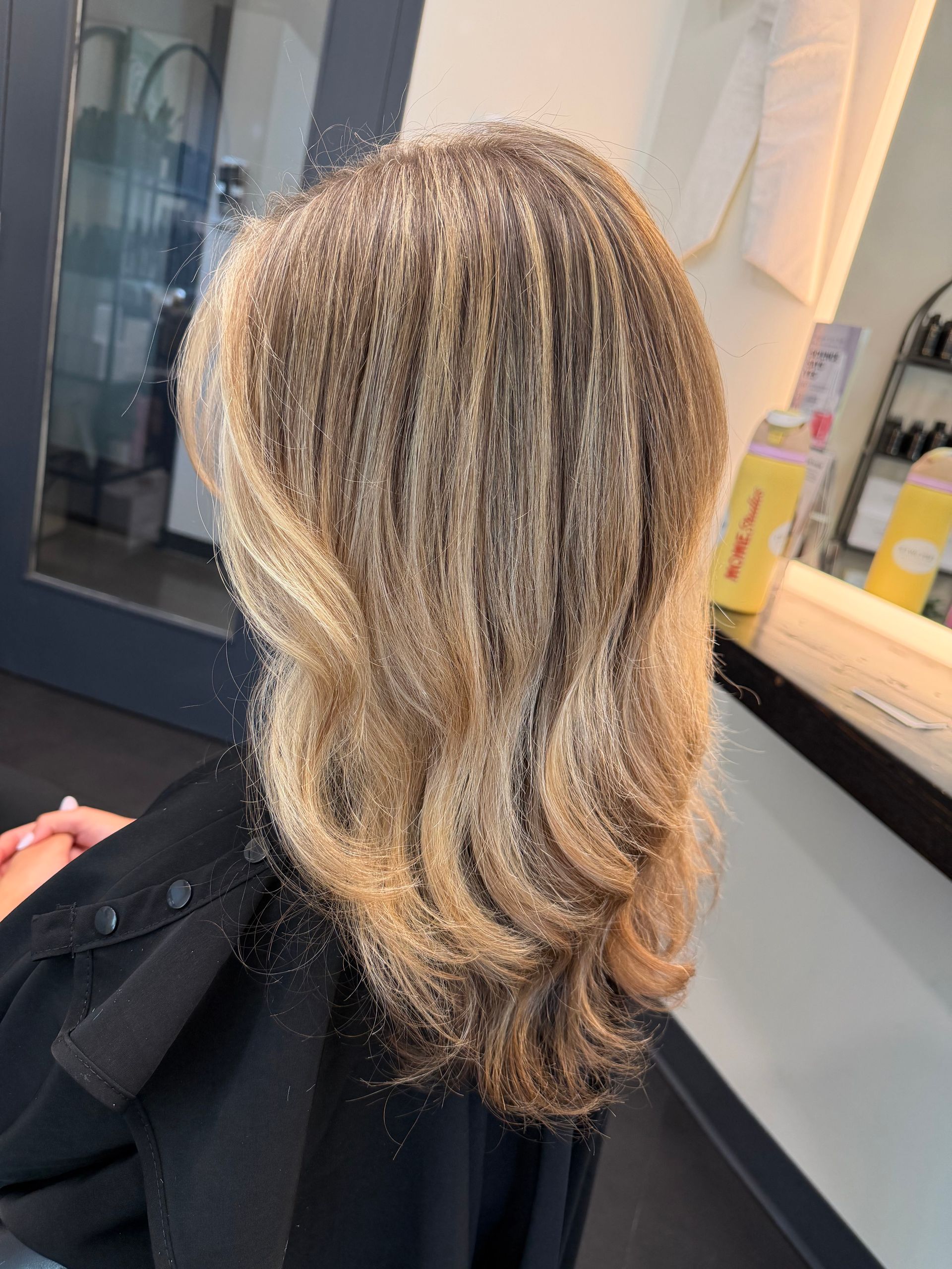 Curly blonde hair with dark roots, styled in a salon chair against a mirror and countertop.