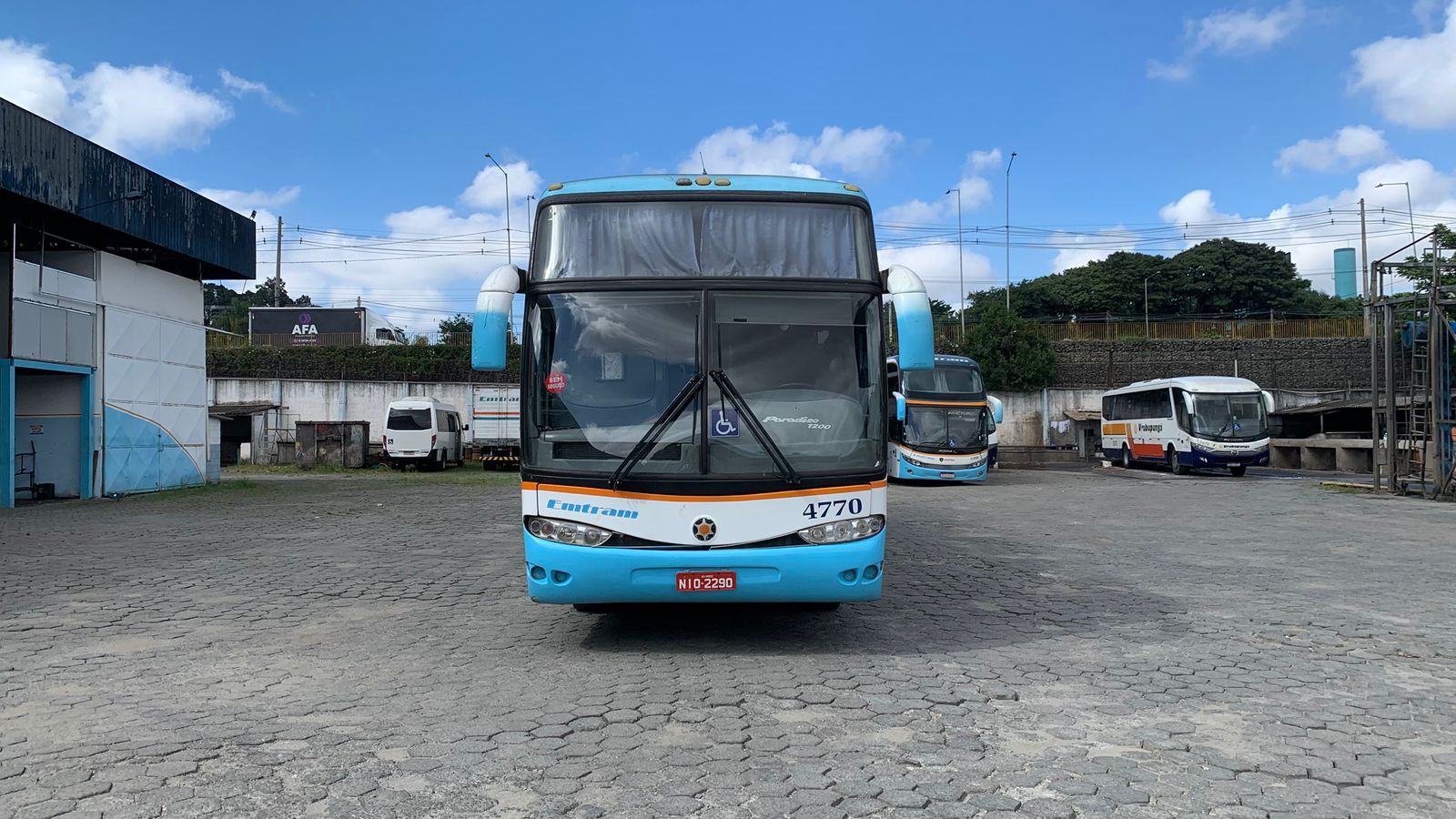 Ônibus azul estacionado em um terreno de cascalho com outros ônibus e um céu nublado ao fundo.