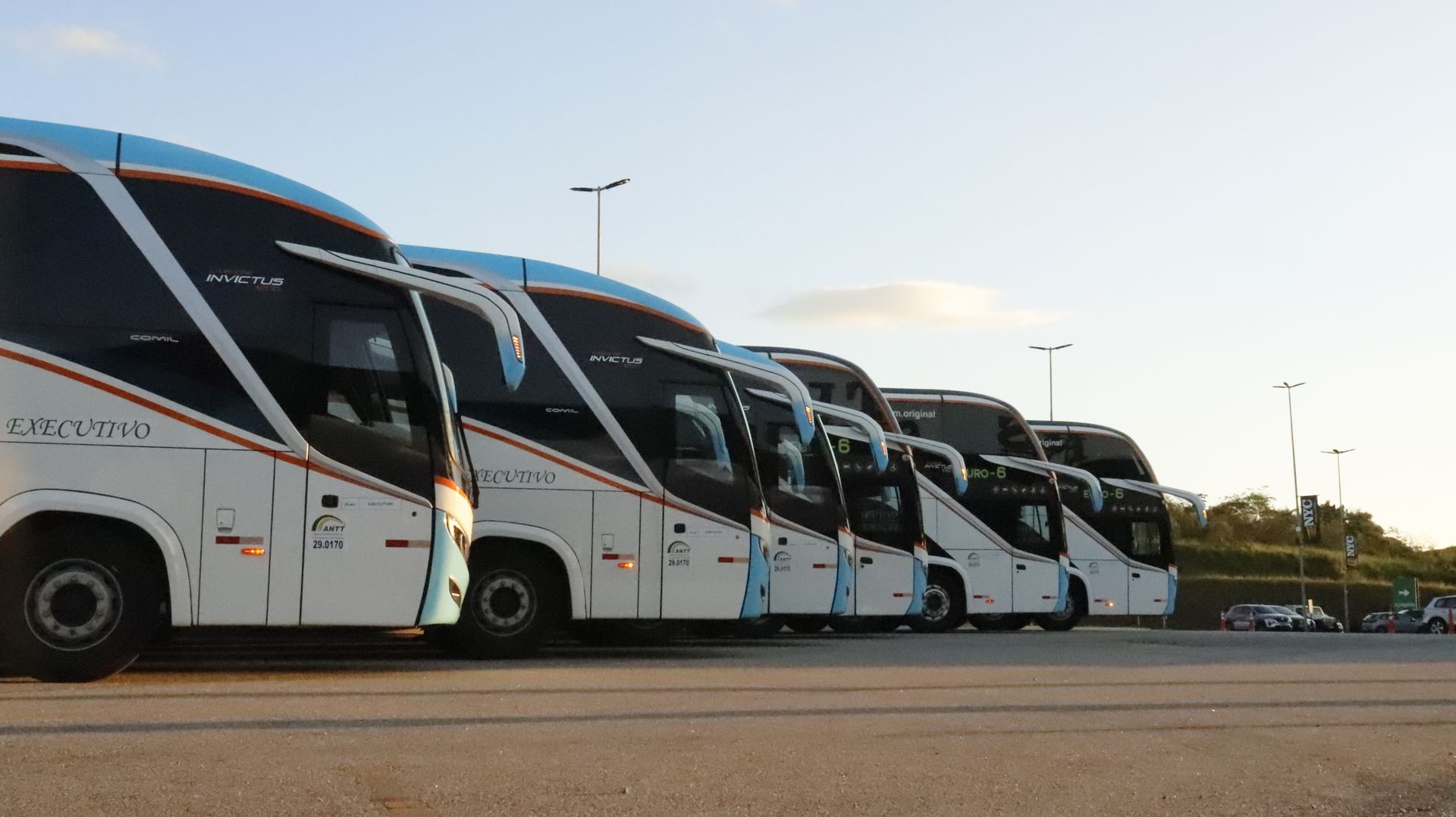 Fileira de ônibus brancos estacionados lado a lado em um estacionamento ao pôr do sol.