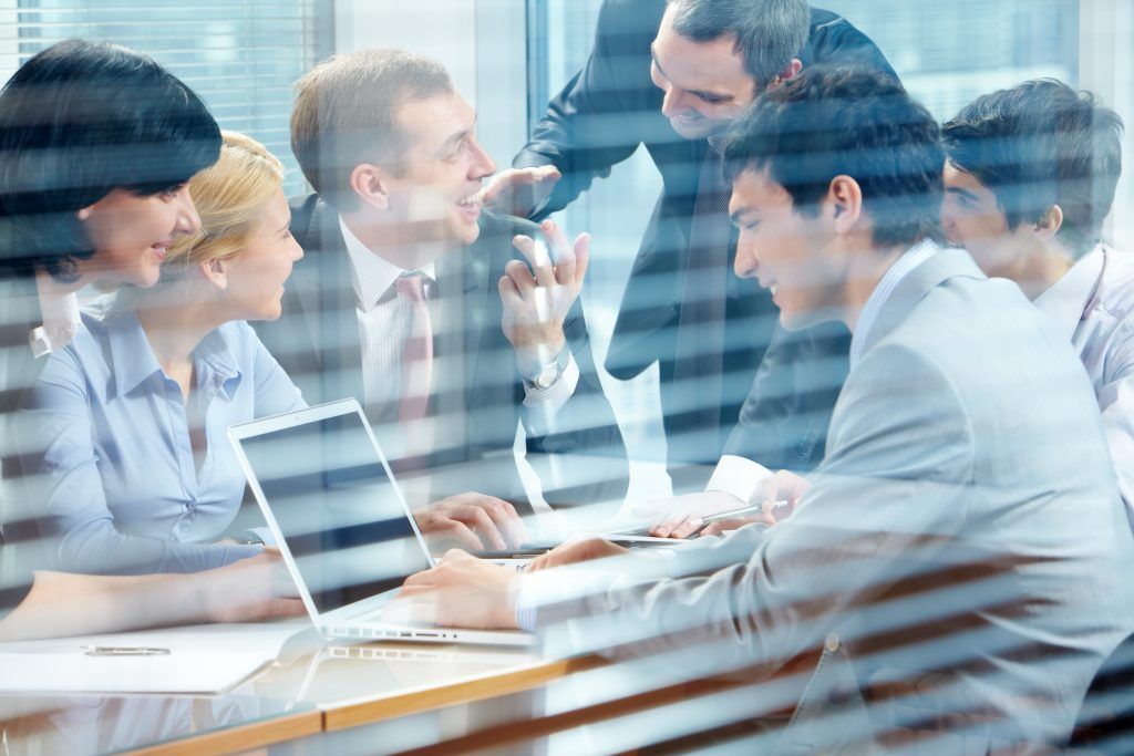 Group of businesspeople in suits collaborating around a laptop at a table, discussing and gesturing.