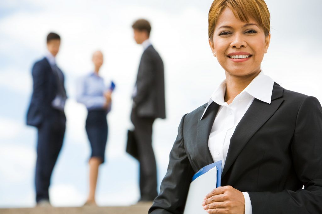Smiling woman in a black suit holds a file; blurred colleagues in background.