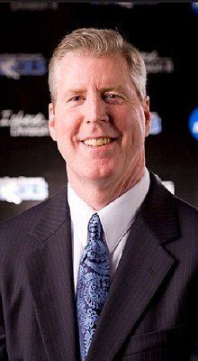 Man in suit smiles at the camera, dark background, tie with blue pattern.