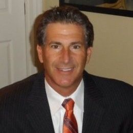 Man in suit, smiling, indoors. He is wearing a dark jacket, white shirt, and orange tie.