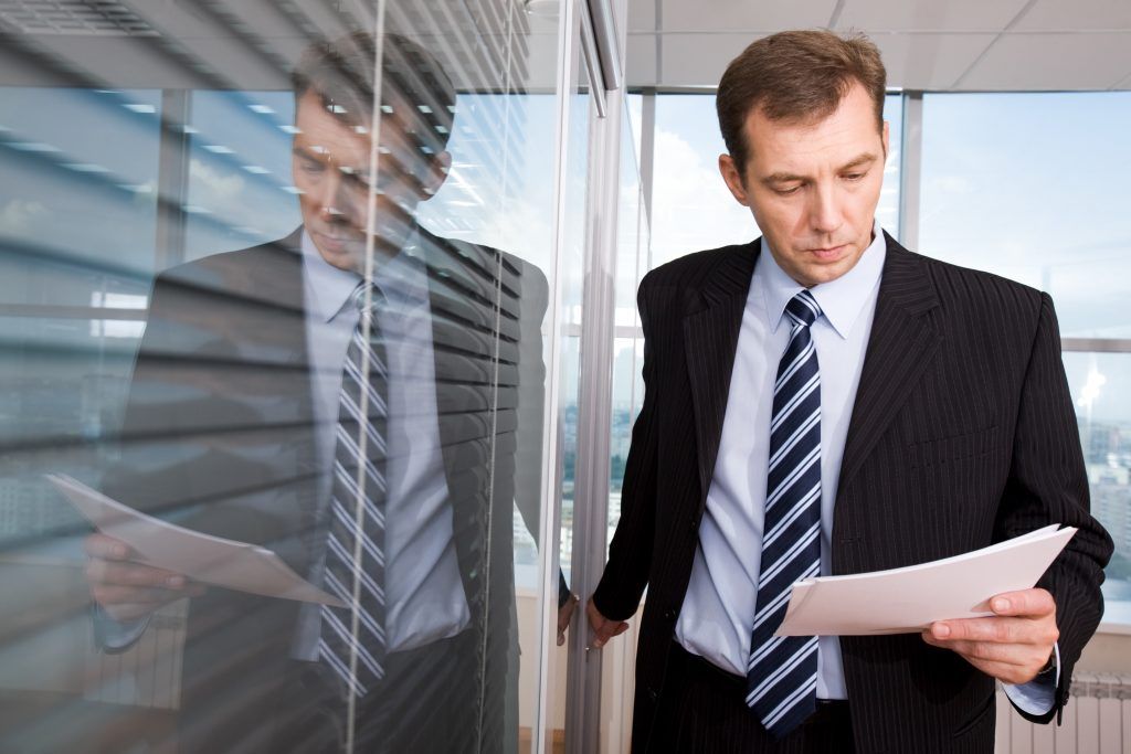Man in suit reading a document near a glass wall with blinds, looking focused.
