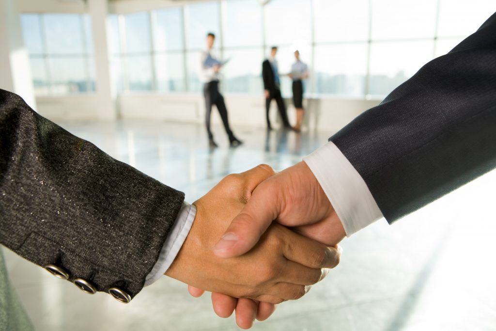 Two business people shaking hands in an office, other people in background.