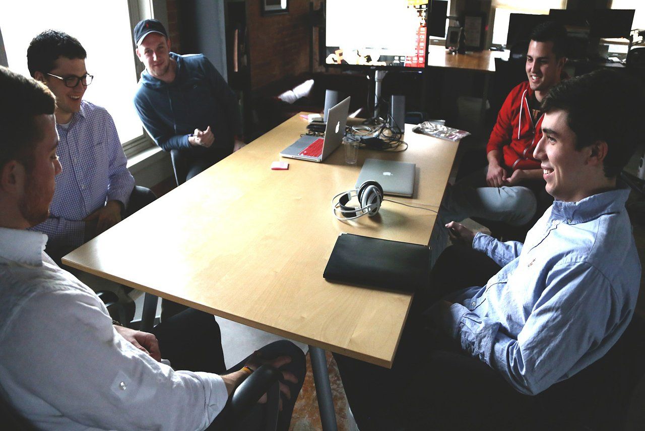 Six men in a meeting around a light wood table, inside a room with a window and a monitor.
