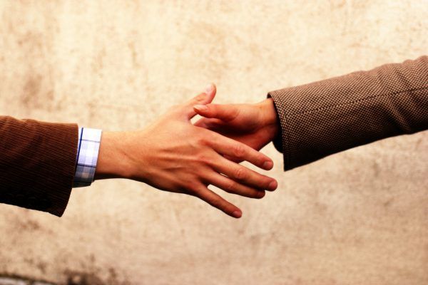 Two people shaking hands against a textured wall, one in a brown jacket, the other in a brown blazer.