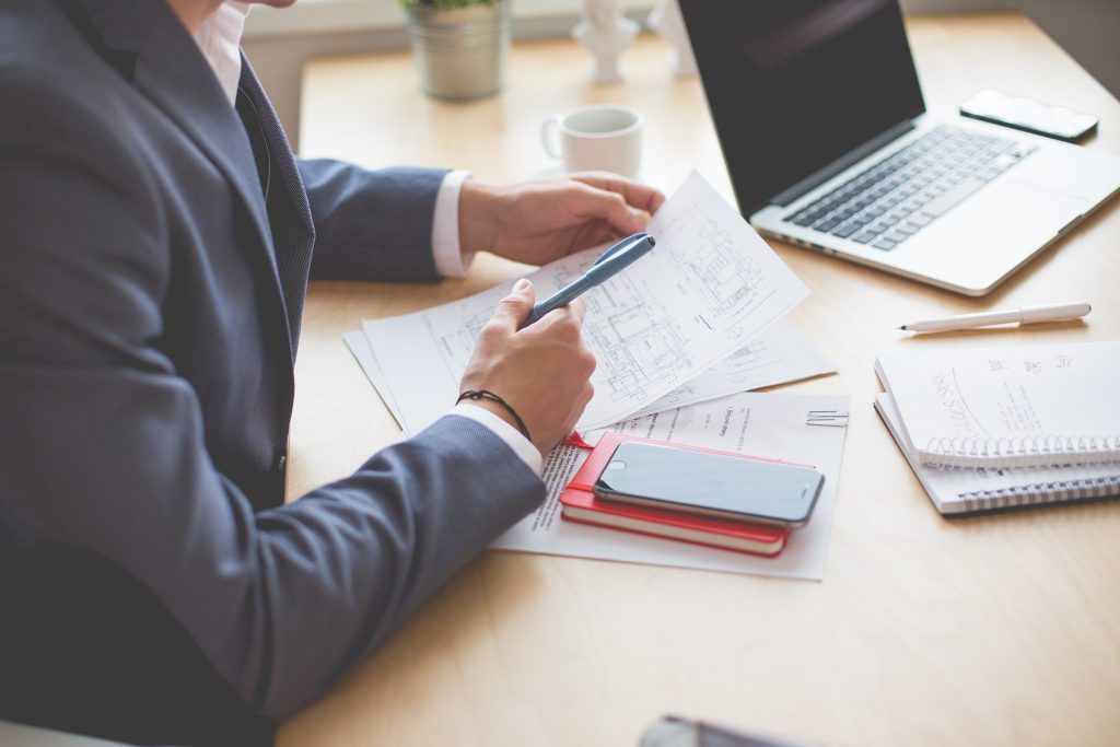 Person in suit reviews documents at a desk with a laptop, phone, and notebook.